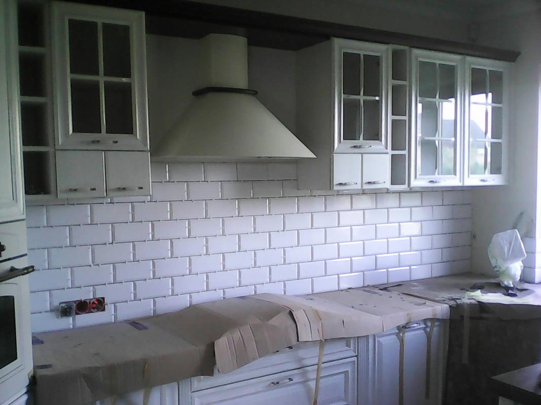 Białe, prostokątne płytki ułożone na ścianie w kuchni, białe szafki wiszące i okap, blat zakryty kartonem ochronnym, widoczne gniazdka elektryczne.