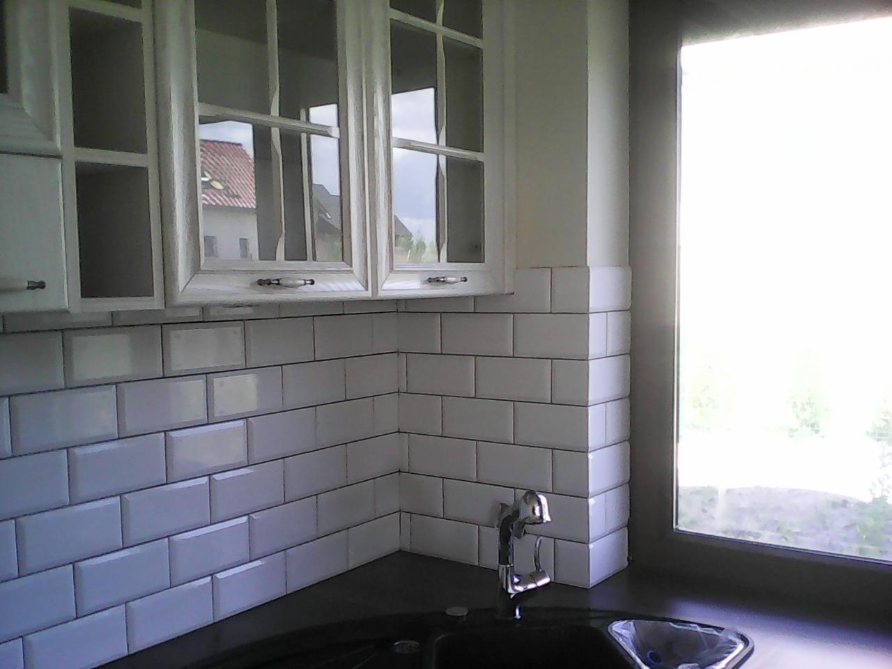 Kuchnia po remoncie: białe, prostokątne płytki ułożone w cegiełkę na ścianie nad blatem, chromowana bateria przy zlewie, białe szafki wiszące z przeszklonymi frontami, widok z okna.