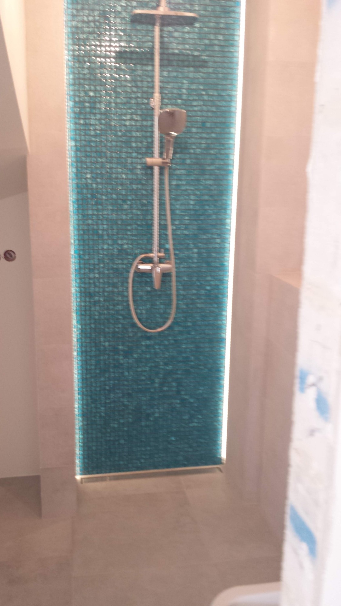 Wykończony prysznic z turkusową mozaiką na ścianie, chromowaną armaturą i beżowymi płytkami podłogowymi.