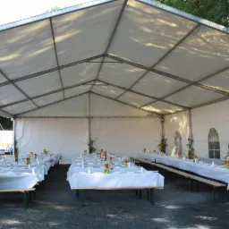 Ustawione stoły pod białym namiotem cateringowym z dekoracjami roślinnymi i zastawą, widoczna obsługa w odblaskowych kamizelkach.