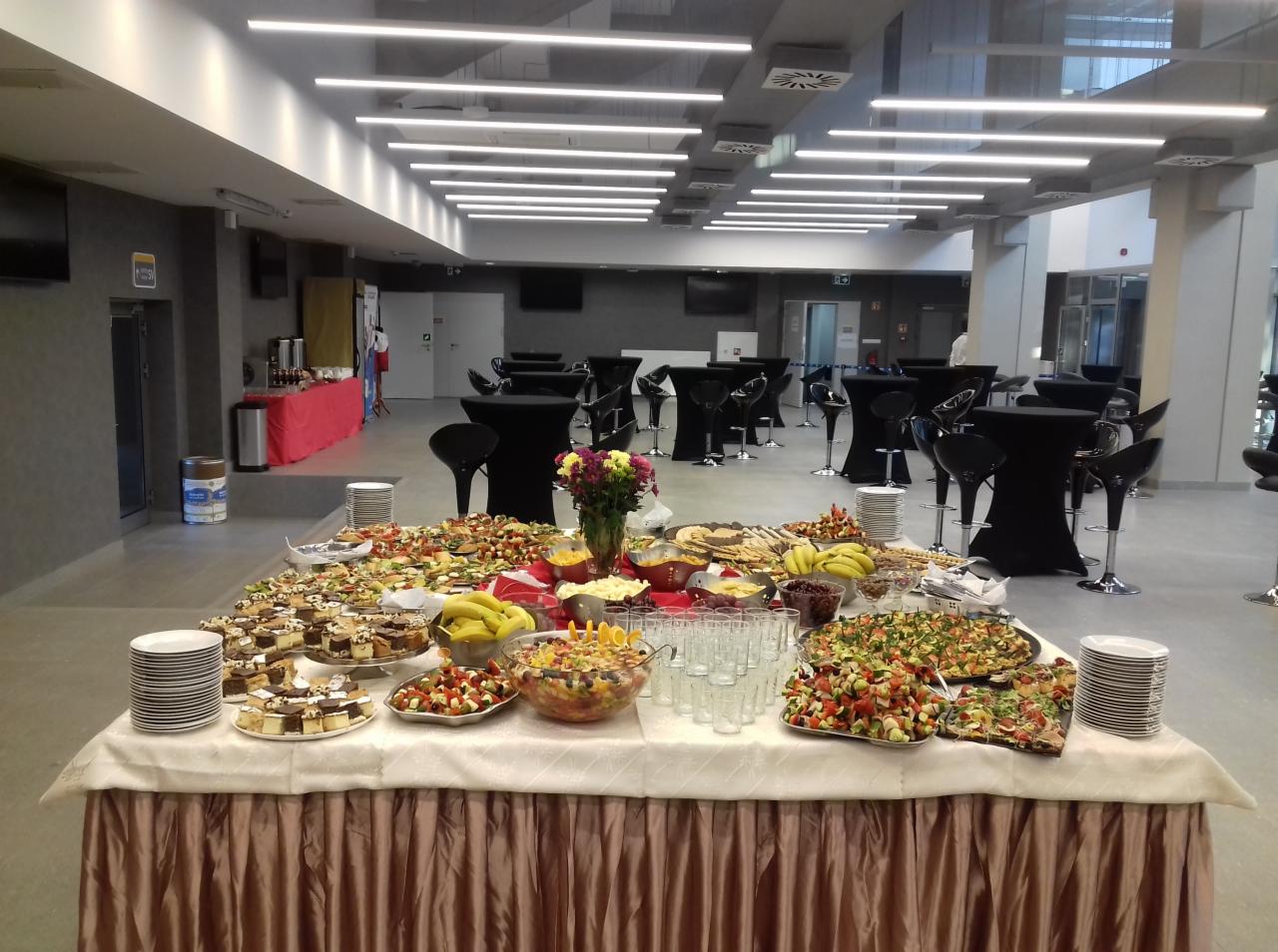 Stół cateringowy z różnorodnymi przekąskami, owocami i deserami w przestronnym, nowoczesnym wnętrzu z czarnymi stolikami barowymi w tle.