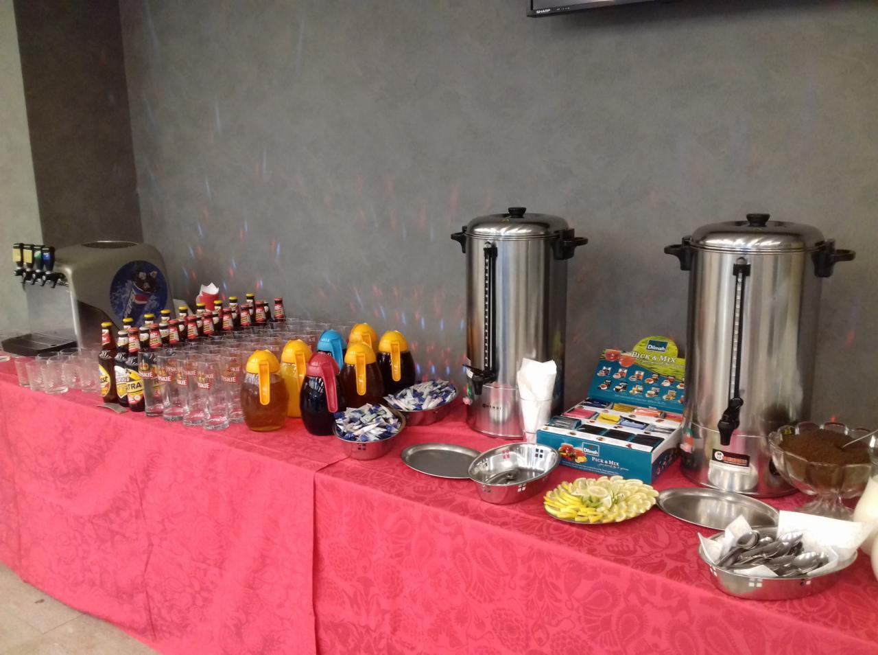 Stół cateringowy nakryty różowym obrusem z przygotowanym napojem z dystrybutora, szklankami, miodem w słoikach, napojami w butelkach, dwoma dużymi termosami, cytryną, cukrem i herbatą Dilmah Pick&Mix.