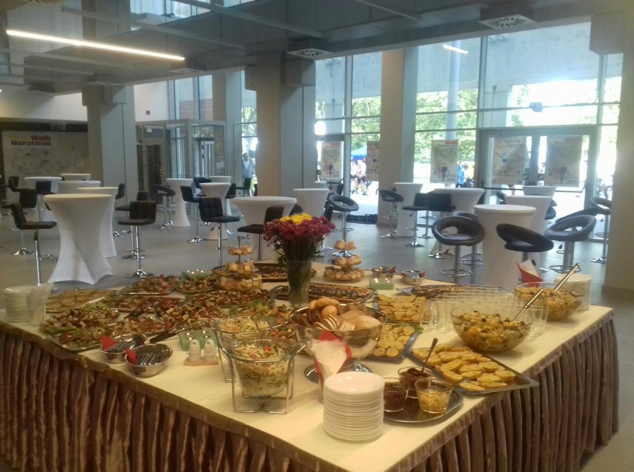 Stół cateringowy z różnorodnymi przekąskami, sałatkami i pieczywem, ustawiony w przestronnym, jasnym wnętrzu z wysokimi oknami i stolikami koktajlowymi w tle.