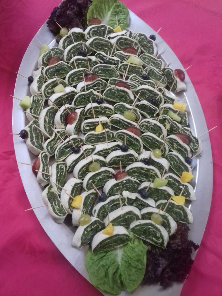 Owalny półmisek z roladkami szpinakowymi, spiętymi wykałaczkami z winogronami i kawałkami mango, dekorowany sałatą na różowym tle.