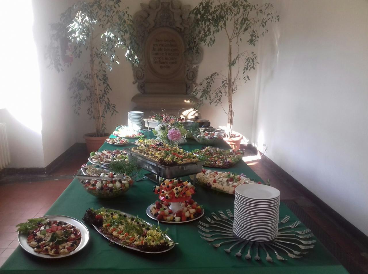 Stół cateringowy z różnorodnymi sałatkami, przekąskami i deserem owocowym w kształcie grzyba, udekorowany kwiatami i otoczony roślinami doniczkowymi w jasnym wnętrzu.