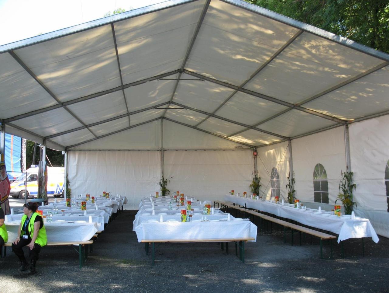 Ustawione stoły pod białym namiotem cateringowym z dekoracjami roślinnymi i zastawą, widoczna obsługa w odblaskowych kamizelkach.