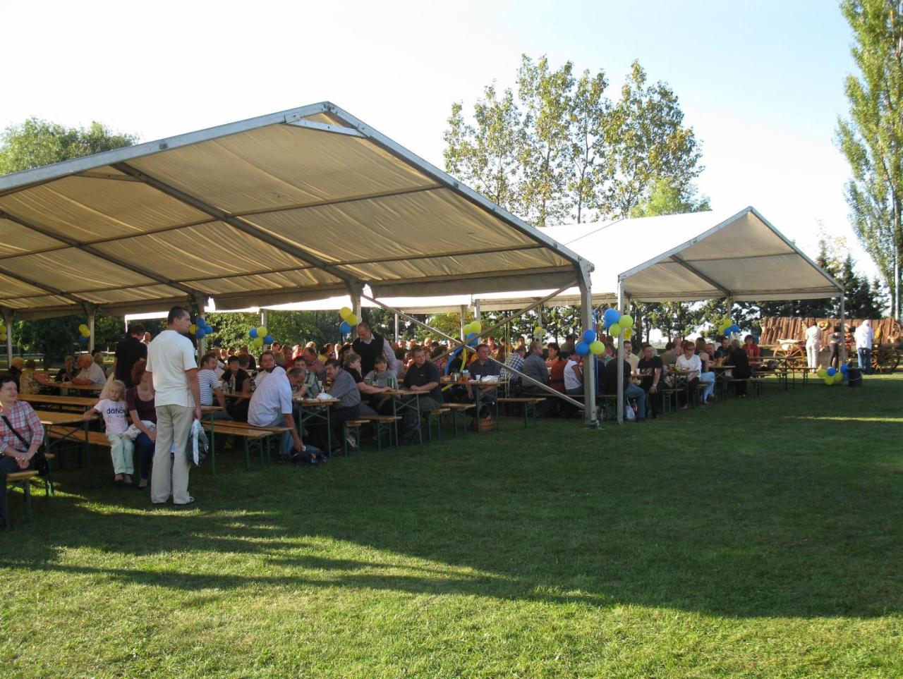 Plenerowa impreza firmowa w Kłodzku pod białymi namiotami, z drewnianymi stołami i ławkami, udekorowana niebieskimi i żółtymi balonami, z licznymi gośćmi.
