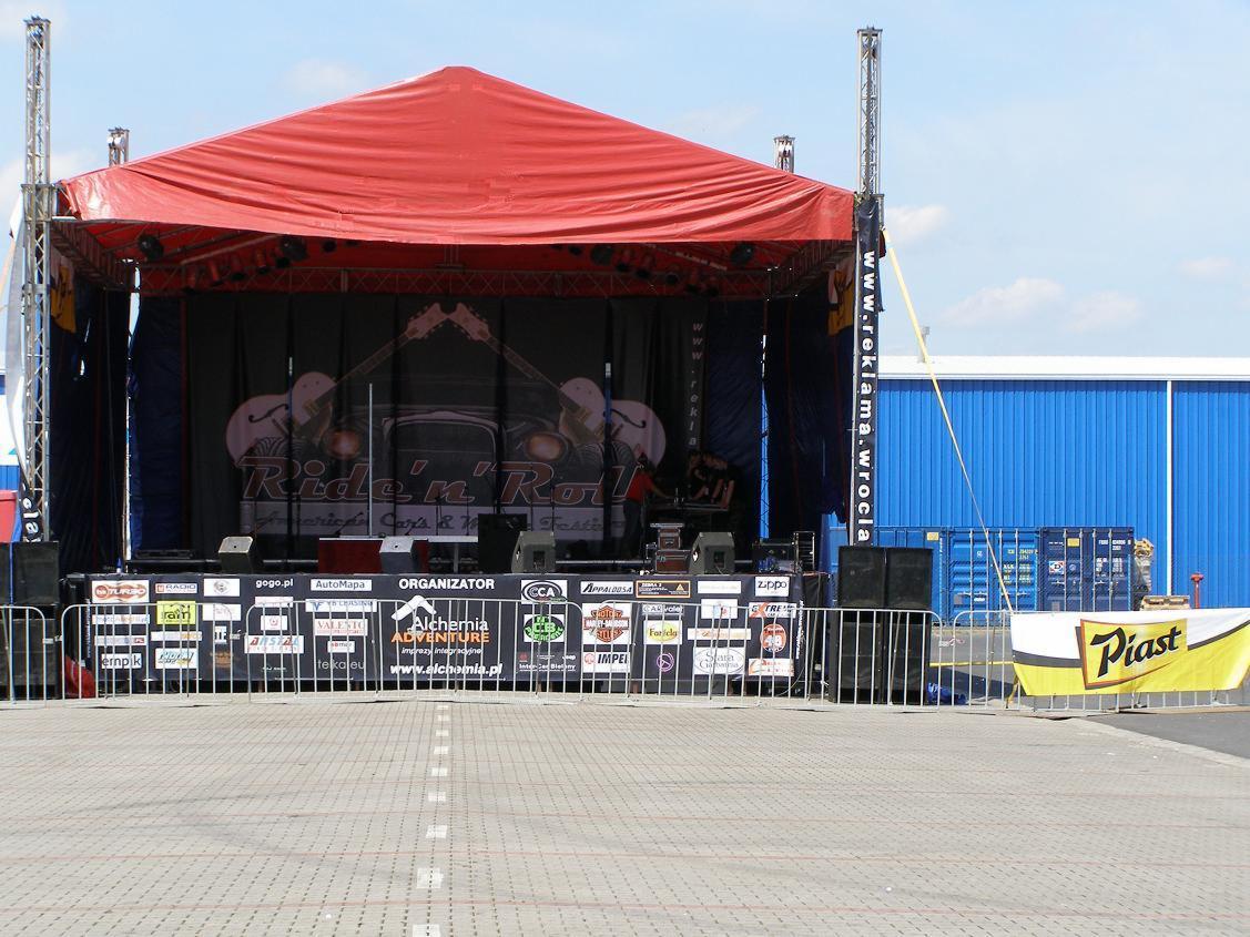 Scena plenerowa z czerwonym zadaszeniem, przygotowana na wydarzenie muzyczne 'Ride'n'Roll', z banerem reklamowym organizatora Alchemia Adventure i sponsorów, zabezpieczona barierkami.