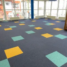 Wykładzina dywanowa w biurze z wzorem kwadratów w kolorach żółtym i miętowym, duże okna z widokiem na miasto, niebieskie okrągłe lampy sufitowe.