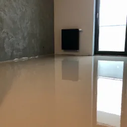 Wnętrze pokoju z wylewką samopoziomującą na podłodze, ściana w szarym dekoracyjnym tynku, czarny grzejnik i okno z widokiem na zewnątrz.