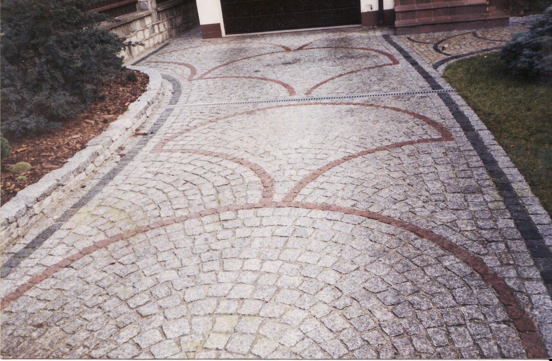 Granitowa kostka brukowa ułożona w łukowaty wzór na podjeździe, z czerwonymi akcentami i odwodnieniem liniowym, prowadząca do garażu.