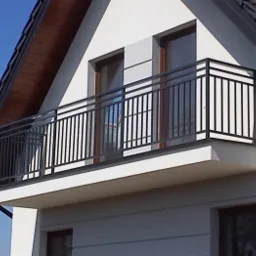 Czarna, metalowa balustrada balkonowa na tle białej elewacji domu jednorodzinnego z drewnianą podbitką dachu i ciemną dachówką.