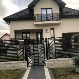 Nowoczesny dom z czarną, metalową bramą i ogrodzeniem o dekoracyjnym wzorze, uzupełniony balkonem z prostą, metalową barierką.