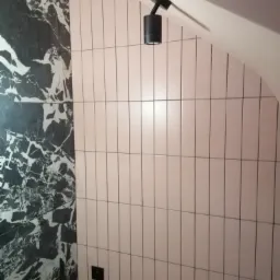 Wykończona ściana łazienki z połączeniem płytek imitujących marmur i podłużnych, różowych płytek ceramicznych, oświetlona czarnym reflektorem sufitowym, widoczny fragment drabiny malarskiej.