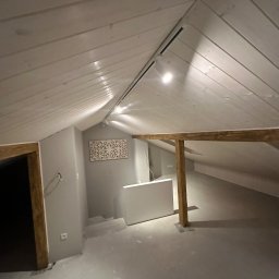 Rafbud - Poddasze po remoncie z drewnianą konstrukcją stropu, białą boazerią na skosach, szarą ścianą z dekoracją i gładką, jasną podłogą przygotowaną do wykończenia.