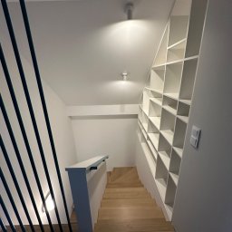 Rafbud - Jasne drewniane schody z czarną balustradą i zabudowaną białą biblioteczką pod skosem na ścianie, oświetlone punktowymi lampami sufitowymi.