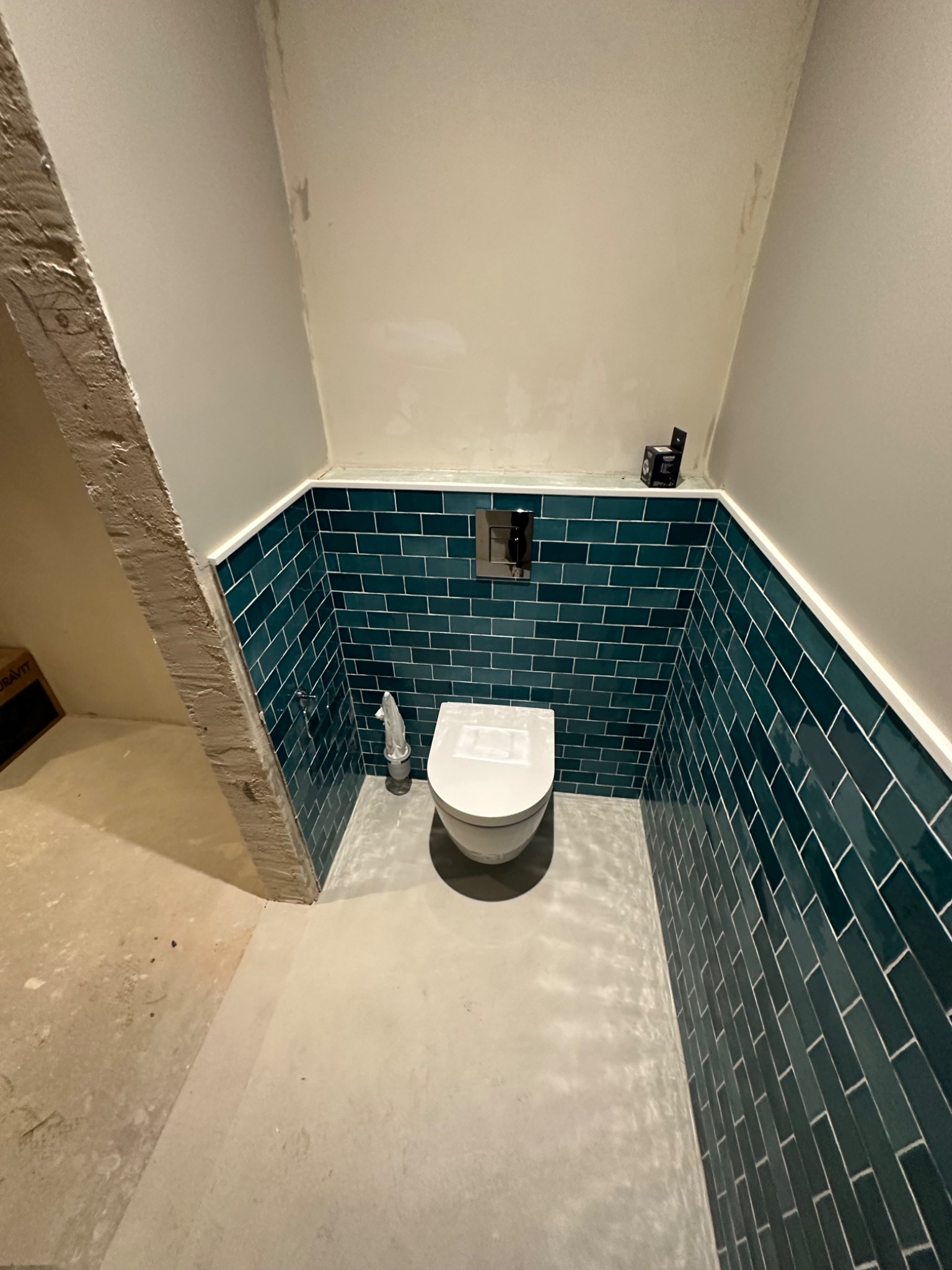 Wnętrze małej łazienki w trakcie prac wykończeniowych, widoczne turkusowe płytki ułożone do połowy wysokości ścian, biała toaleta i surowy betonowy filar z lewej strony.