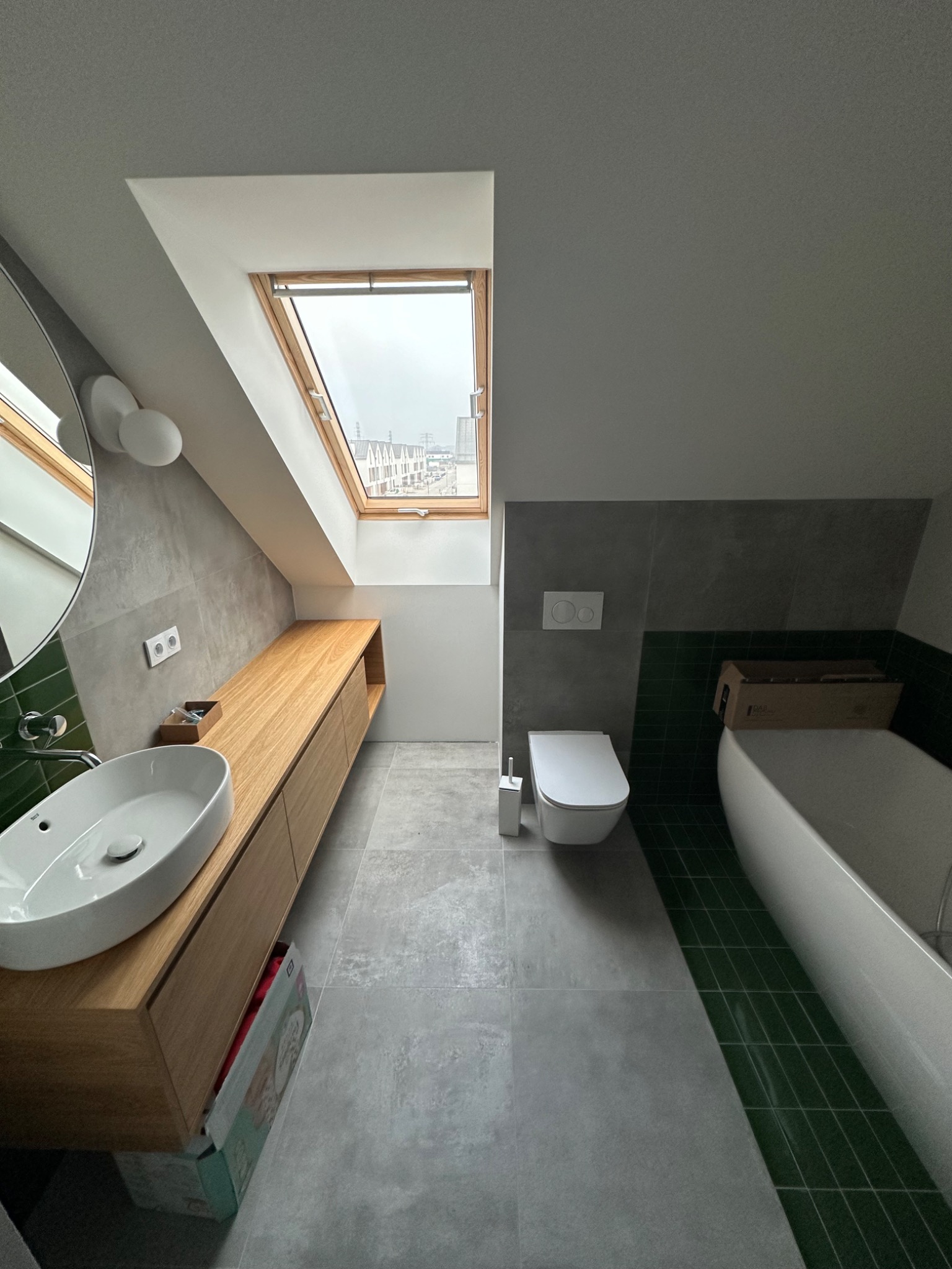 Nowoczesna łazienka na poddaszu ze skośnym oknem dachowym, drewnianą szafką pod umywalką, szarą płytką na ścianach i podłodze oraz zielonym akcentem z płytek przy wannie.