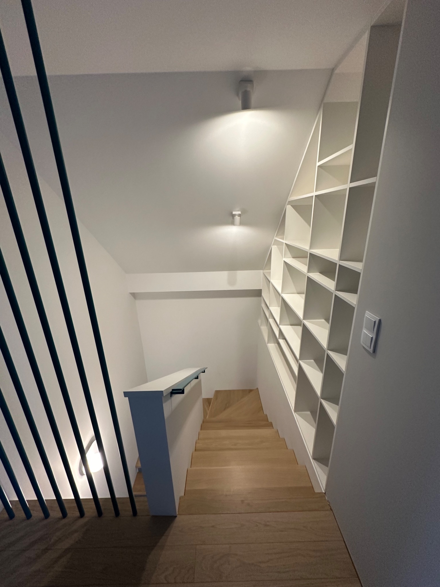 Jasne drewniane schody z czarną balustradą i zabudowaną białą biblioteczką pod skosem na ścianie, oświetlone punktowymi lampami sufitowymi.