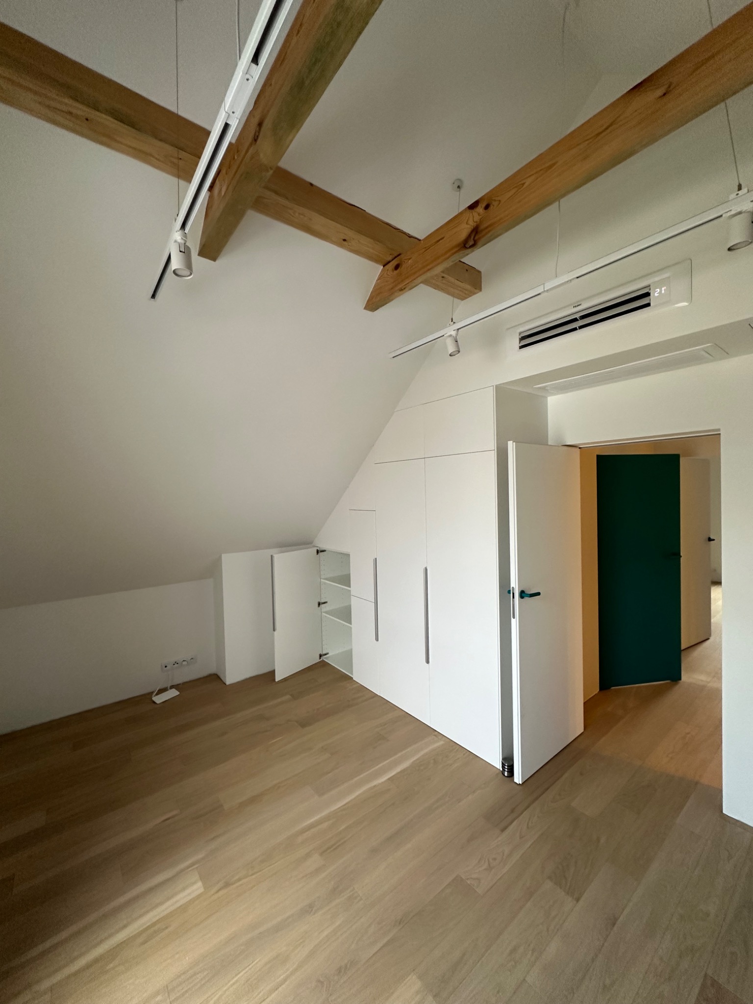 Poddasze z drewnianymi belkami, białymi szafami w zabudowie i parkietem, widoczne gniazdka elektryczne przy podłodze i klimatyzator pod sufitem.