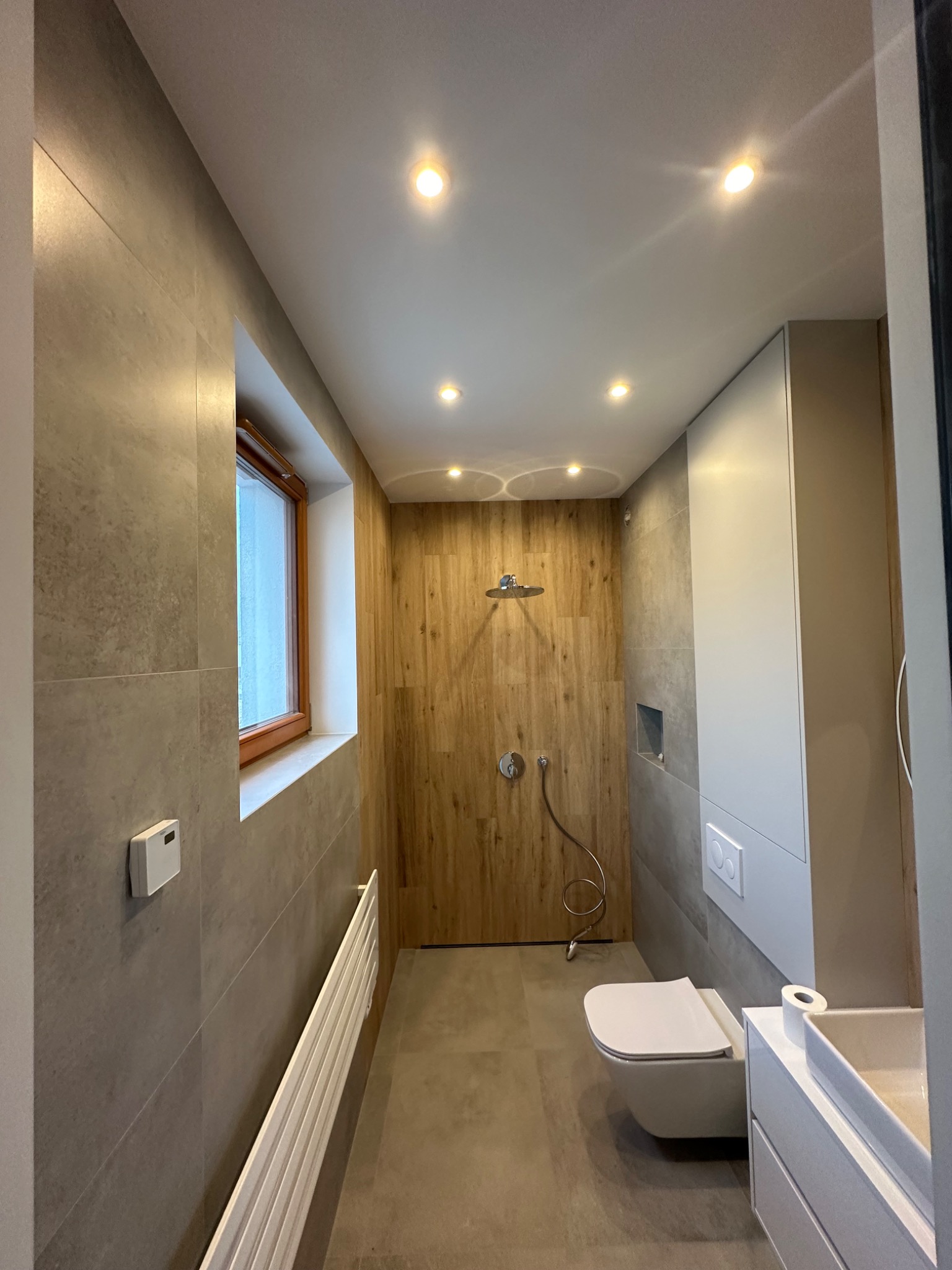 Nowoczesna łazienka z drewnianą ścianą prysznicową, szarą ceramiką, podtynkową toaletą i białymi meblami łazienkowymi podświetlona punktowymi lampami sufitowymi.