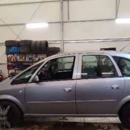 Srebrny samochód typu hatchback na stanowisku do geometrii kół w warsztacie, z widocznymi urządzeniami pomiarowymi przy kołach i monitorem z wynikami pomiarów.