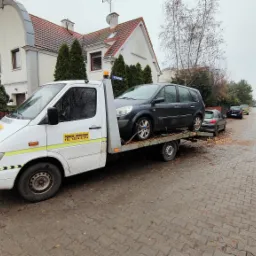 Biała laweta z załadowanym szarym samochodem osobowym na tle brukowanej drogi i budynków mieszkalnych w jesiennej scenerii.