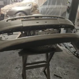 Pomalowany na złoto-beżowy kolor, zdemontowany przedni zderzak samochodowy, ustawiony na drewnianym stołku w warsztacie samochodowym. W tle samochód zabezpieczony folią malarską i narzędzia...