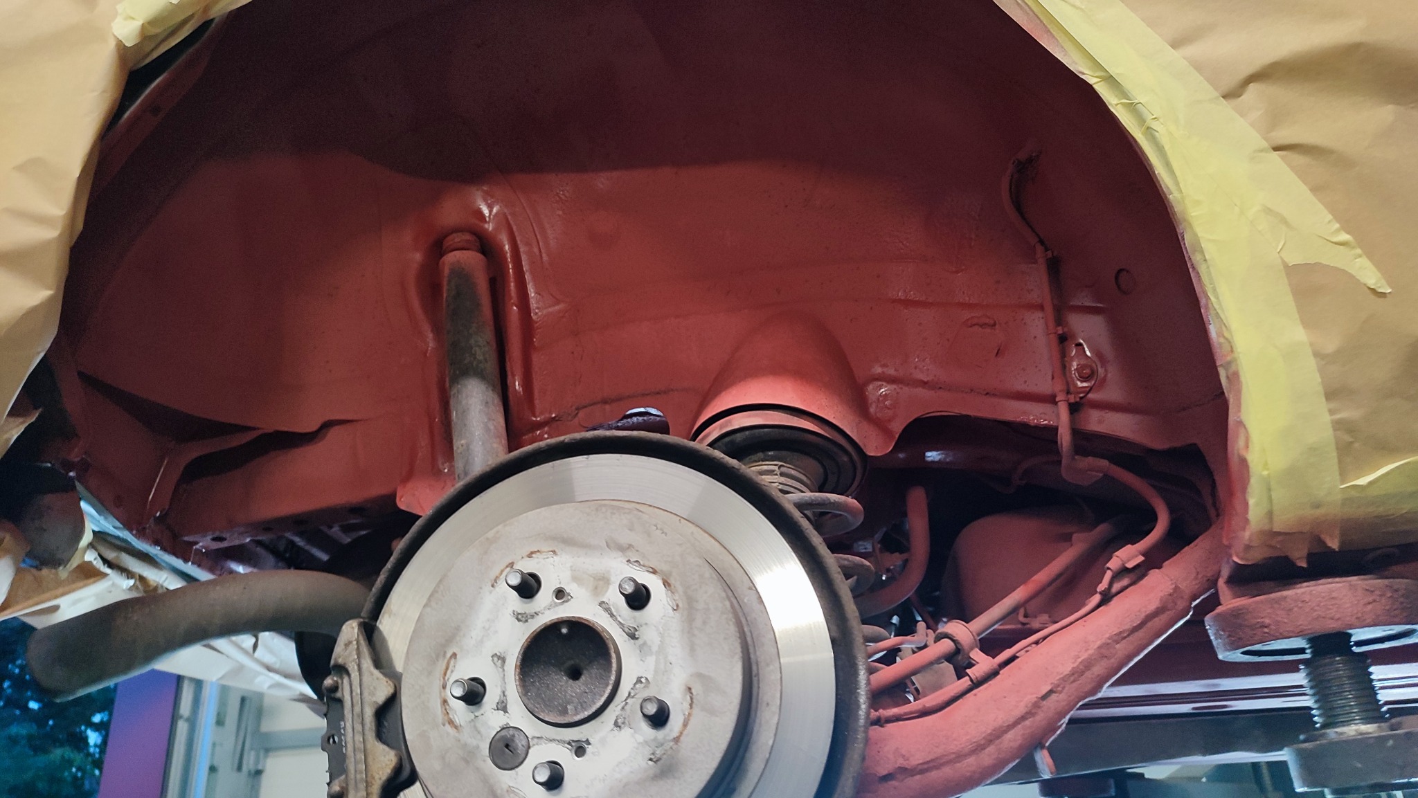 Wnętrze nadkola samochodu po nałożeniu czerwonej powłoki antykorozyjnej, widoczne elementy zawieszenia, tarcza hamulcowa i maskowanie papierem.