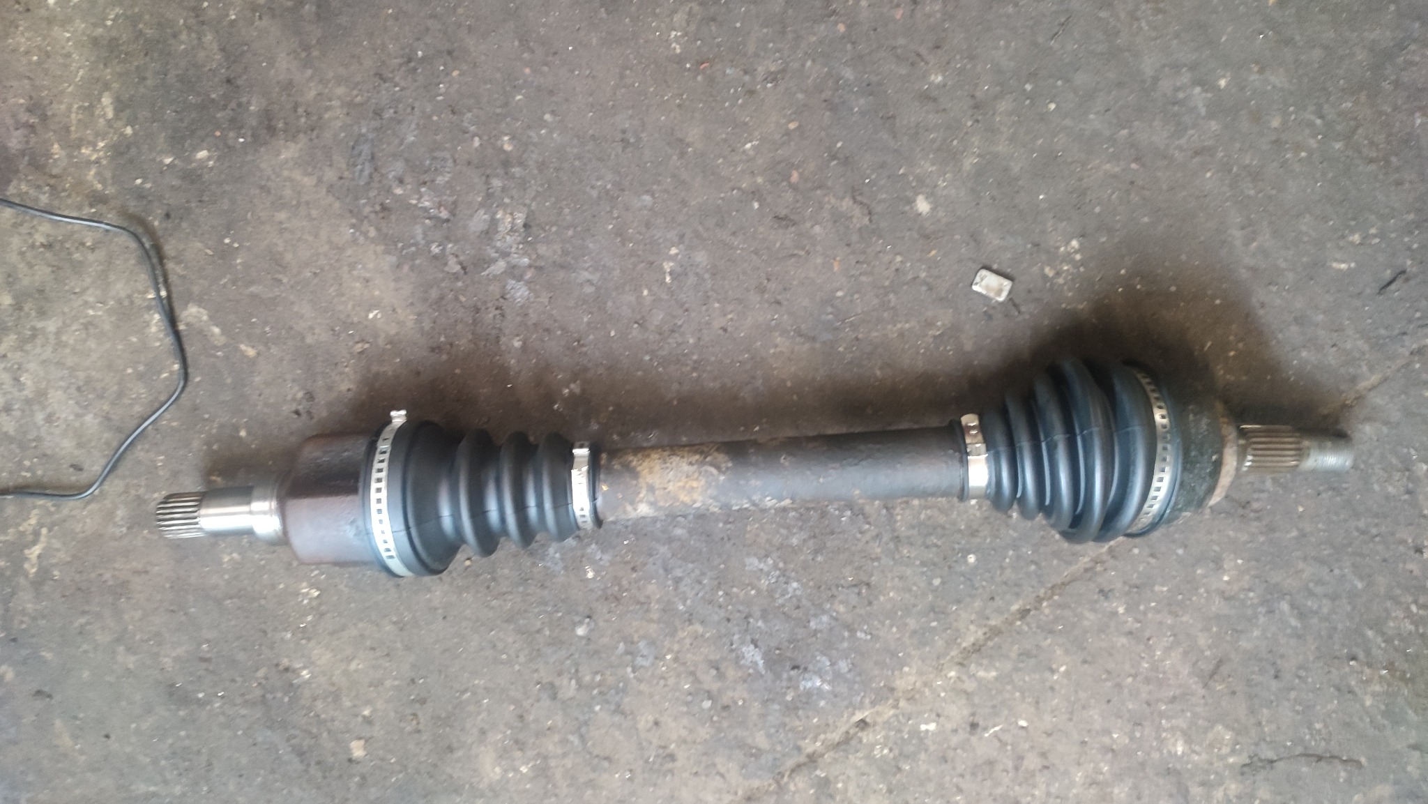 Używana półoś napędowa z przegubami, leżąca na betonowej posadzce warsztatu, widoczne ślady korozji i zużycia.