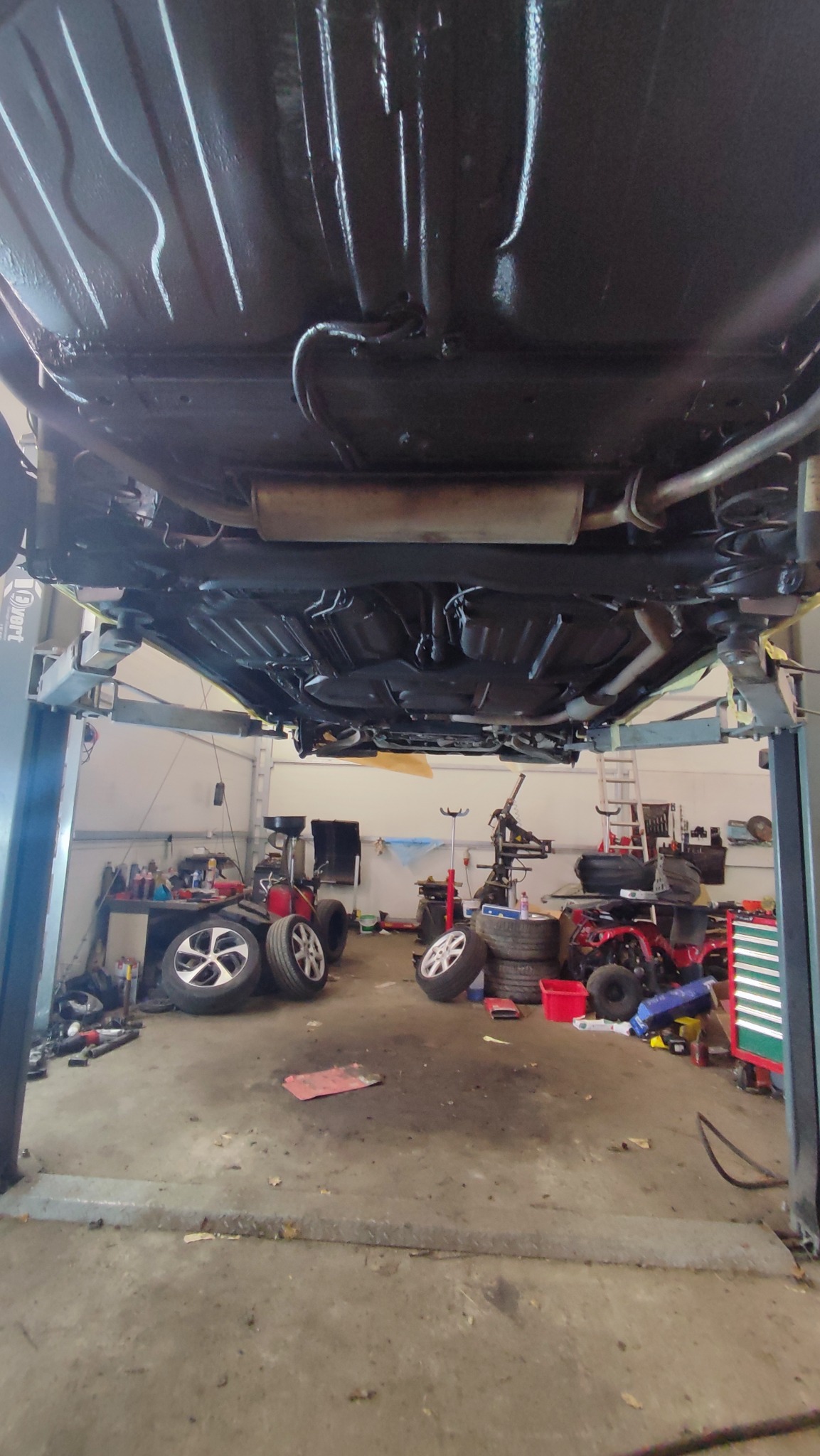 Podwozie samochodu po zabezpieczeniu antykorozyjnym, widoczne na podnośniku w warsztacie, z elementami układu wydechowego i zawieszenia pokrytymi czarną powłoką ochronną.