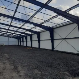 Wnętrze hali w trakcie budowy, widoczna stalowa konstrukcja nośna, ściany z jasnych płyt warstwowych, podłoże gruntowe z gruzem, widok w głąb hali.