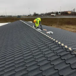 Pracownik w żółtym ubraniu roboczym montuje elementy na czarnym dachu z dachówki ceramicznej, w tle widoczna droga szybkiego ruchu z samochodami.