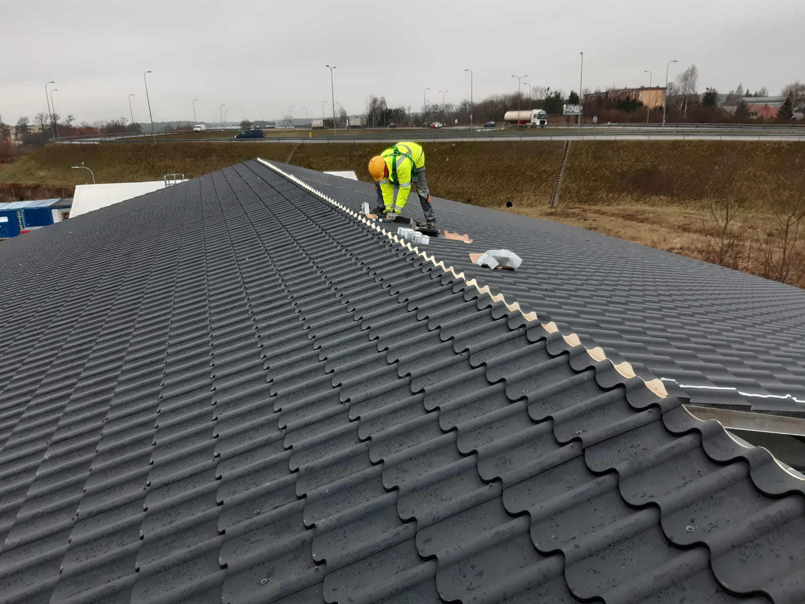 Pracownik w żółtym ubraniu roboczym montuje elementy na czarnym dachu z dachówki ceramicznej, w tle widoczna droga szybkiego ruchu z samochodami.