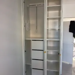 Wnętrze białej, trzydrzwiowej szafy z otwartymi drzwiami, widoczne półki, szuflady i drążek na wieszaki. Szafa stoi na szarej wykładzinie.