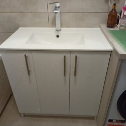 Kamil Tomczak - Biała umywalka z szafką pod spodem i chromowaną baterią w łazience z beżowymi płytkami, obok pralka.