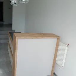 Biała lada recepcyjna z drewnianymi elementami w nowoczesnym wnętrzu z szarą podłogą i wiszącymi lampami kulami.