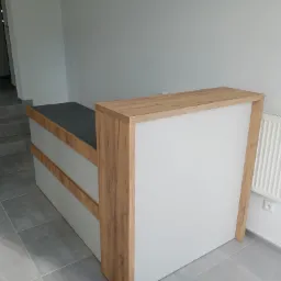 Nowoczesna recepcja biurowa z blatem w kolorze ciemnego grafitu i frontem w odcieniach bieli i drewna, widoczny grzejnik na ścianie w tle.