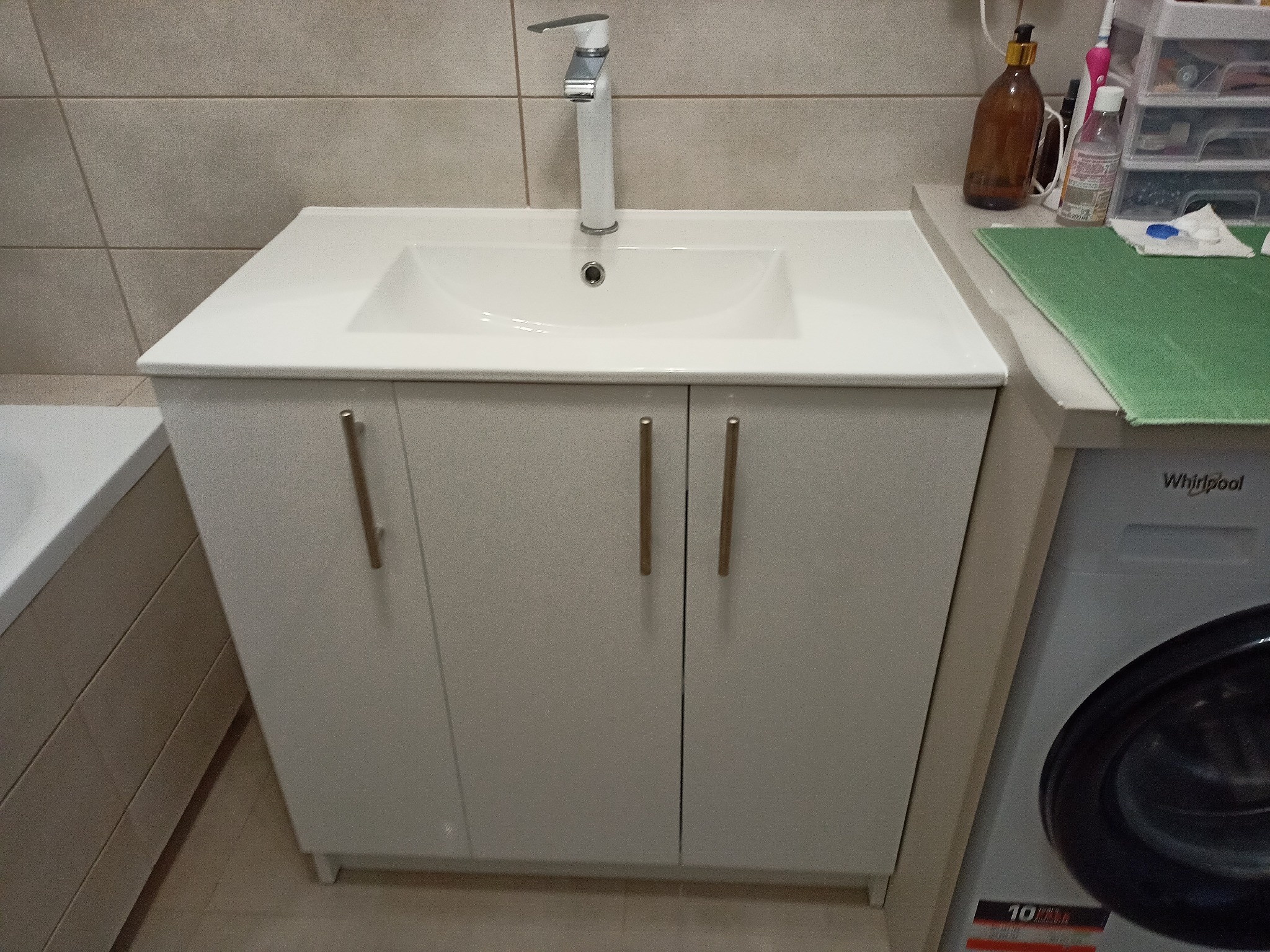 Biała umywalka z szafką pod spodem i chromowaną baterią w łazience z beżowymi płytkami, obok pralka.