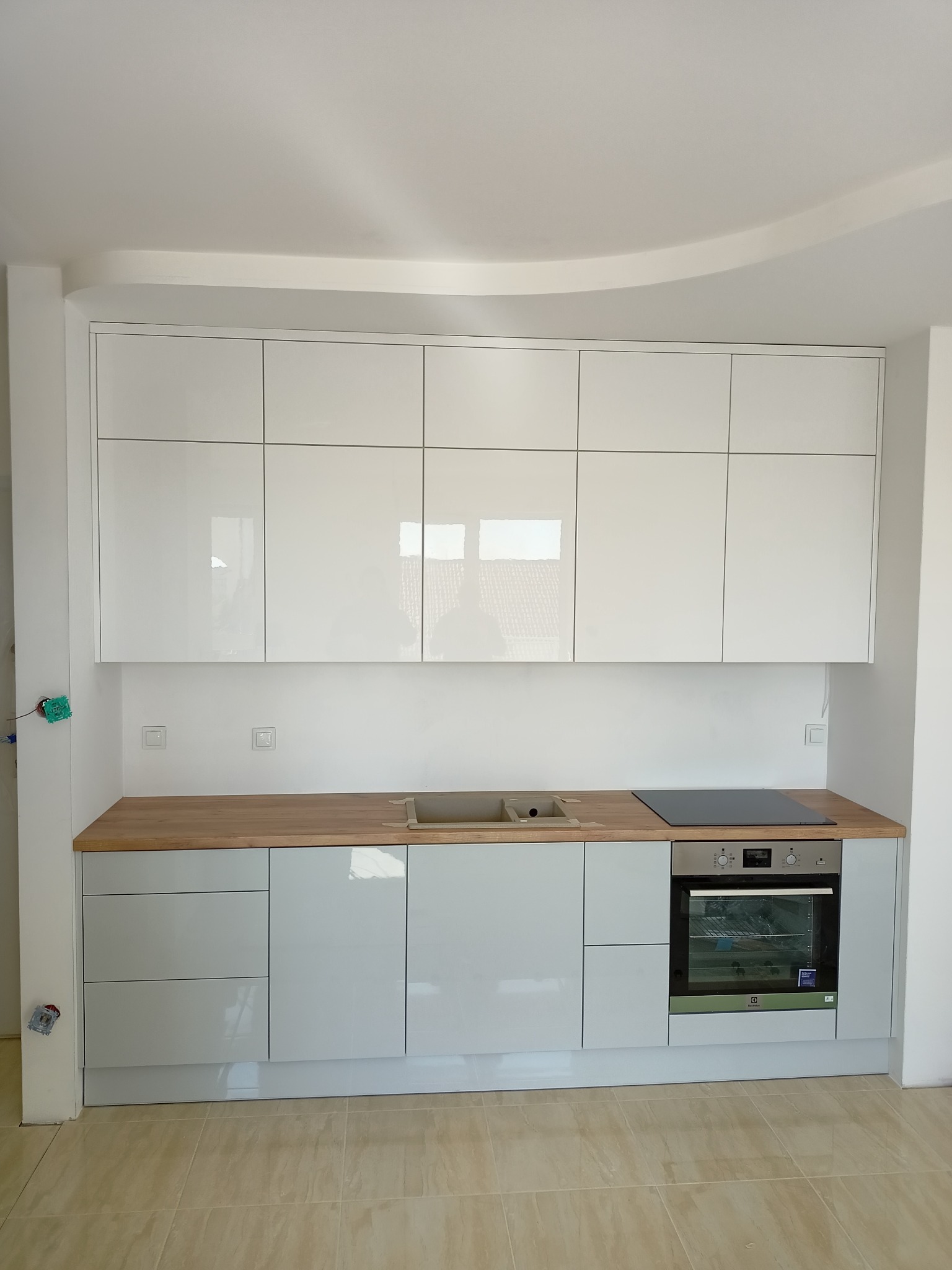 Jasna, nowoczesna kuchnia z białymi błyszczącymi szafkami górnymi i szarymi dolnymi, drewnianym blatem, płytą indukcyjną i piekarnikiem. Widoczne niedokończone prace elektryczne przy ścianie.