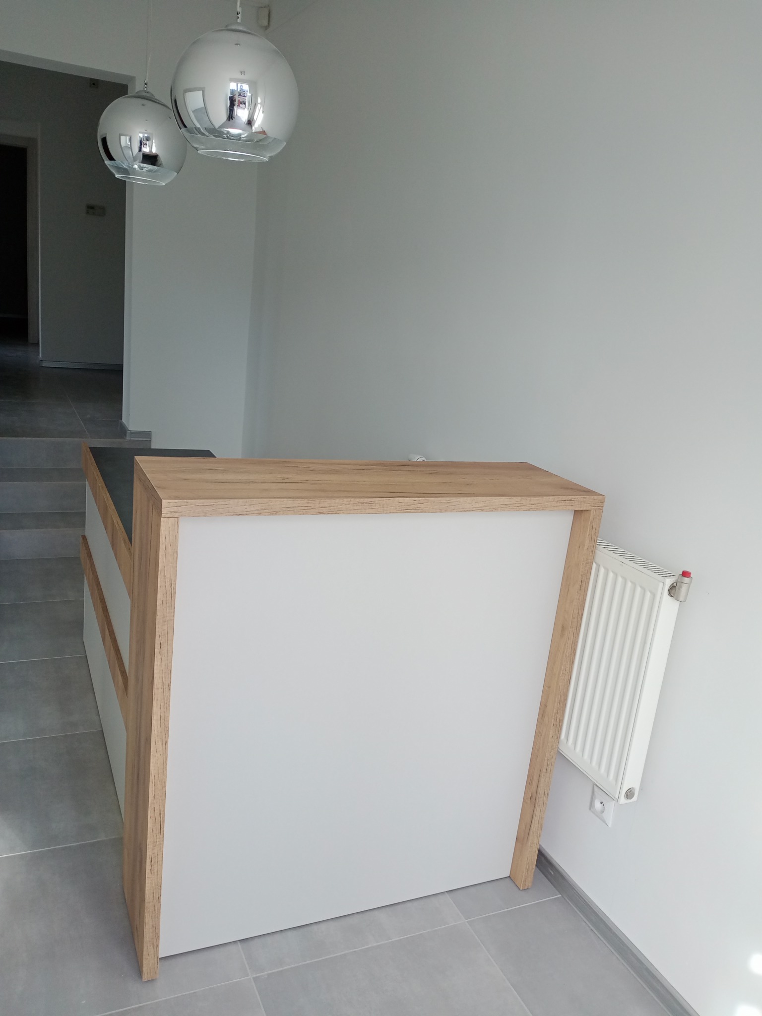 Biała lada recepcyjna z drewnianymi elementami w nowoczesnym wnętrzu z szarą podłogą i wiszącymi lampami kulami.