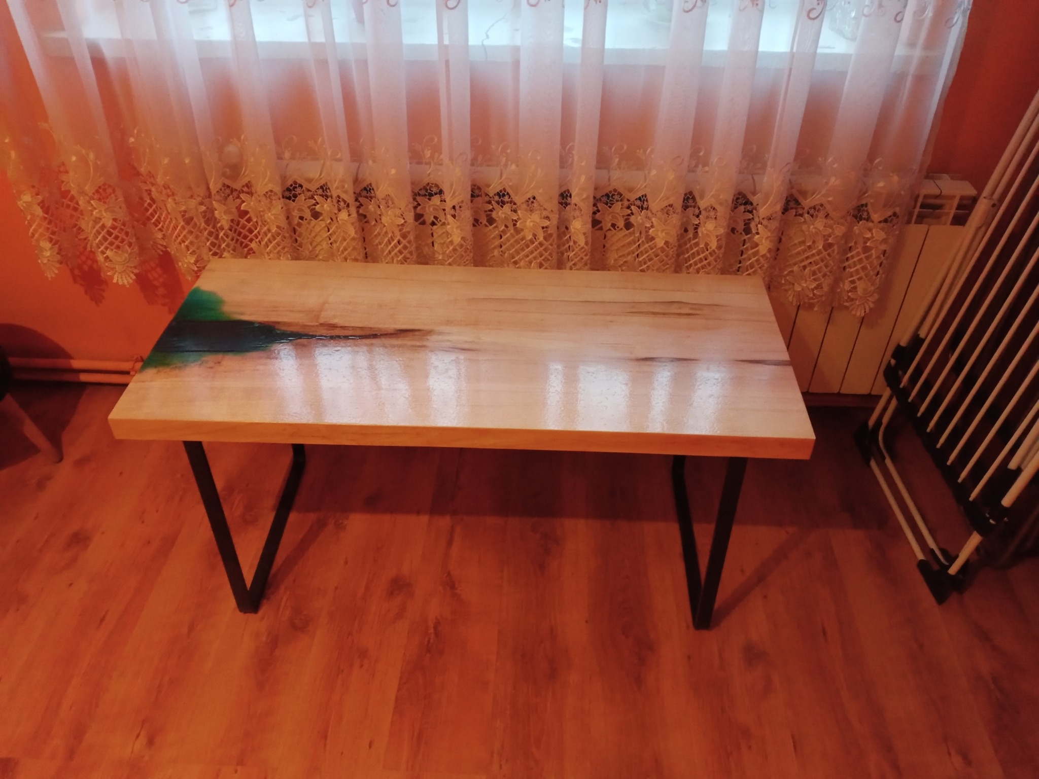 Niski stolik kawowy z blatem z jasnego drewna z żywicą epoksydową w kolorze zielonym, na czarnych, metalowych nogach w kształcie litery V, ustawiony na podłodze w pokoju z pomarańczowymi ścianami...