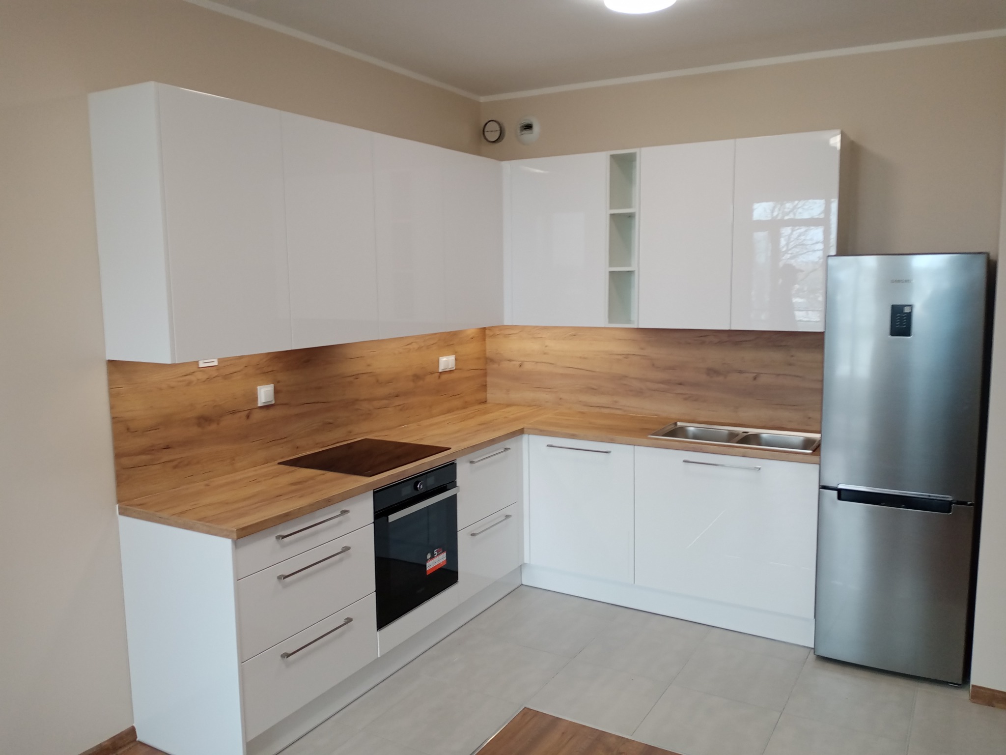 Jasna kuchnia na wymiar z białymi, lakierowanymi frontami i drewnopodobnym blatem oraz srebrną lodówką side-by-side.