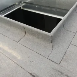 Szczegółowe ujęcie płaskiego dachu pokrytego papą z obróbką blacharską wokół otworu świetlika lub wyłazu dachowego.