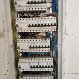 Otwarta skrzynka rozdzielcza z widocznymi bezpiecznikami, przewodami i listwami zaciskowymi, zamontowana na surowej ścianie z betonu komórkowego.