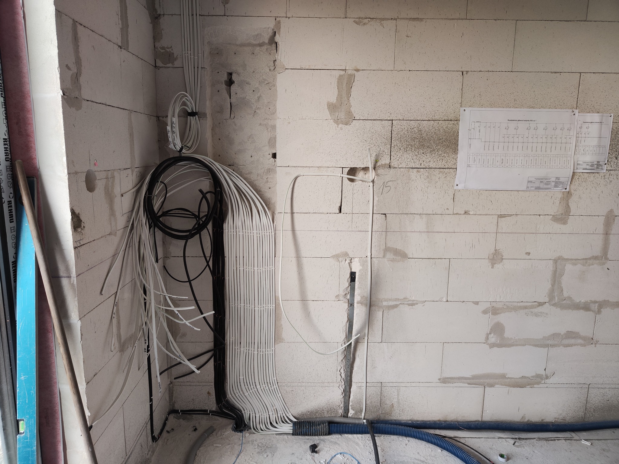 Wiązka kabli elektrycznych, białych i czarnych, prowadzona wzdłuż ściany z betonu komórkowego w surowym stanie, z widocznym schematem elektrycznym na ścianie i niebieskimi rurami karbowanymi...