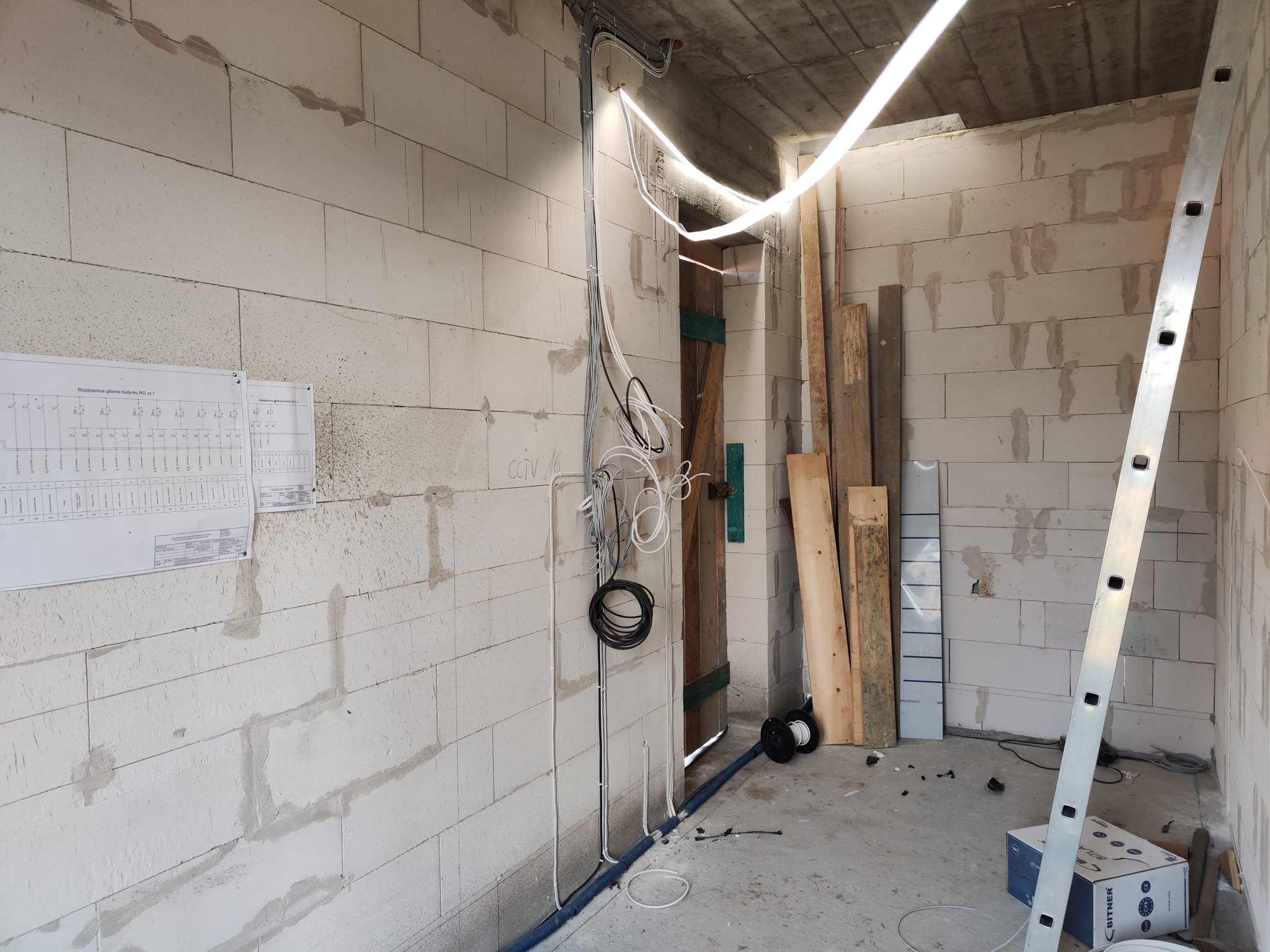 Wnętrze surowego pomieszczenia w trakcie prac elektrycznych: widoczne przewody prowadzone po ścianie z bloczków, aluminiowa drabina oparta o ścianę, rozdzielnica z schematem na ścianie,...