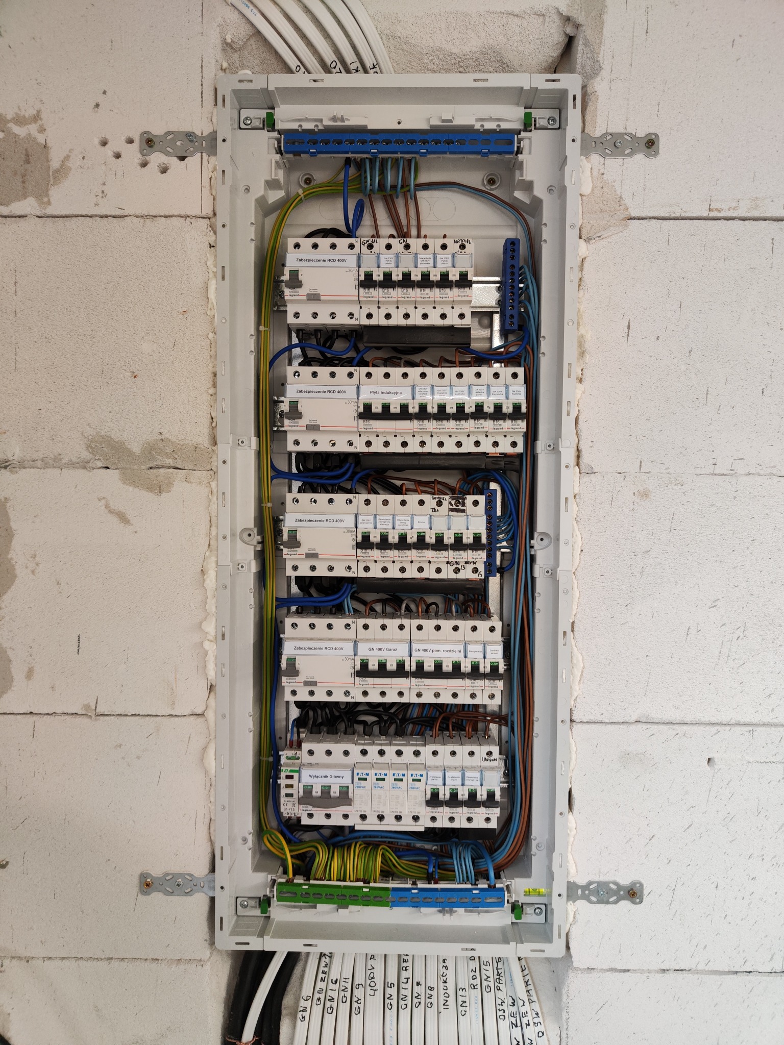 Rozdzielnica elektryczna z zamontowanymi bezpiecznikami i okablowaniem, umieszczona w surowej ścianie z betonu komórkowego, widoczne oznaczenia przewodów.