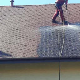 Pracownik w czerwonym kombinezonie, zabezpieczony linami, czyści dach z bitumicznej dachówki za pomocą myjki ciśnieniowej; widoczna różnica w kolorze między umytą a brudną powierzchnią dachu.