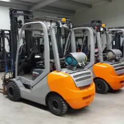 Rząd nowych wózków widłowych marki STILL w magazynie, modele spalinowe z pomarańczową obudową i butlą gazową oraz elektryczne z szarą obudową, widoczne w tle kolejne wózki.